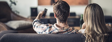 Tampilan belakang pasangan yang duduk di sofa ruang keluarga dan sedang menonton TV, si pria memegang remote