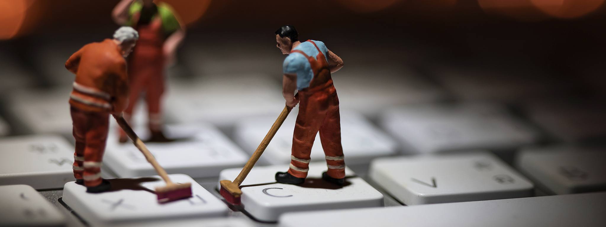 Miniature people cleaning white keyboard computer, with orange lights flickering in the background